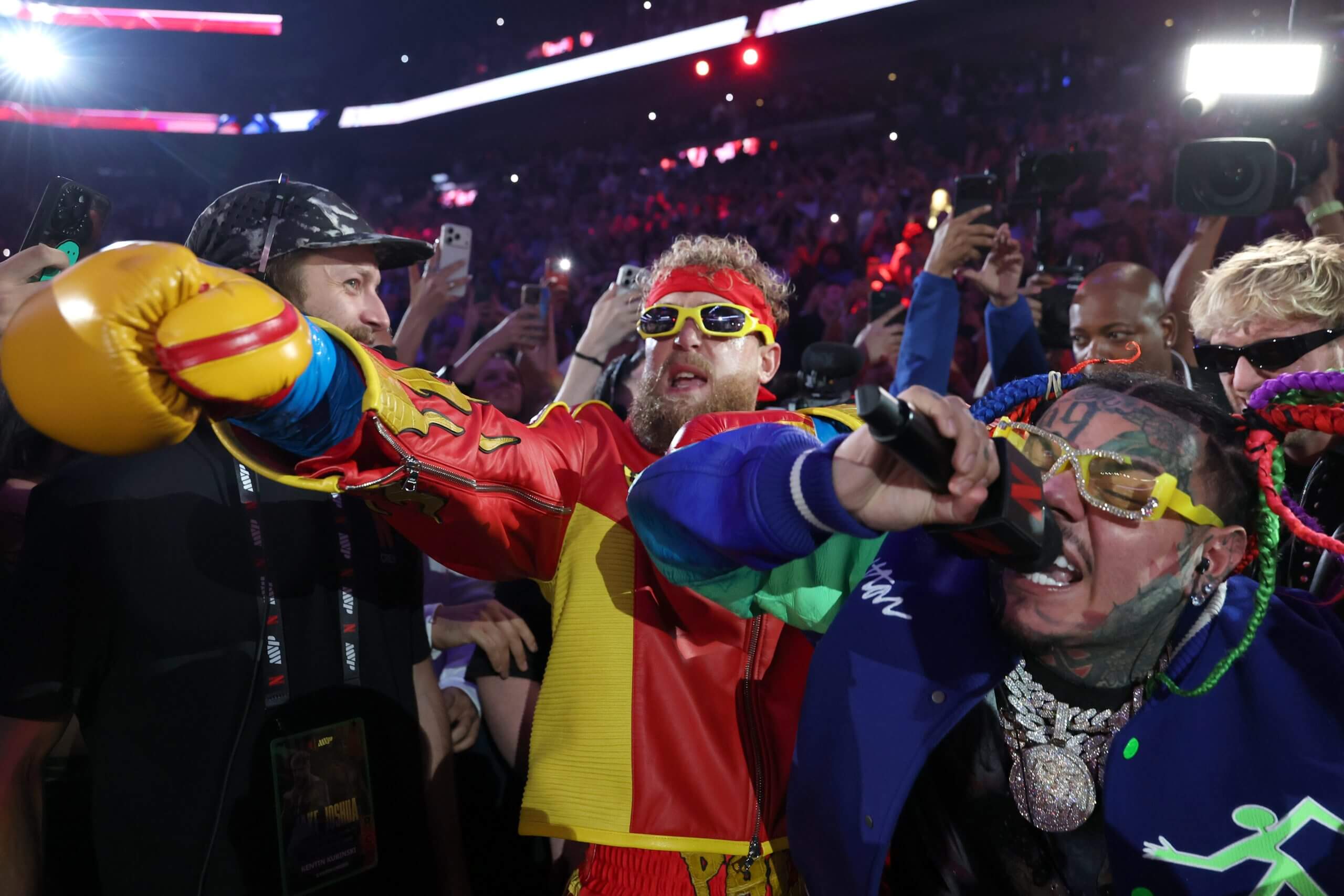 6ix9ine sings as he walks with Jake Paul, wearing yellow sunglasses and a red sweatband, to the ring before his heavyweight bout against Anthony Joshua.