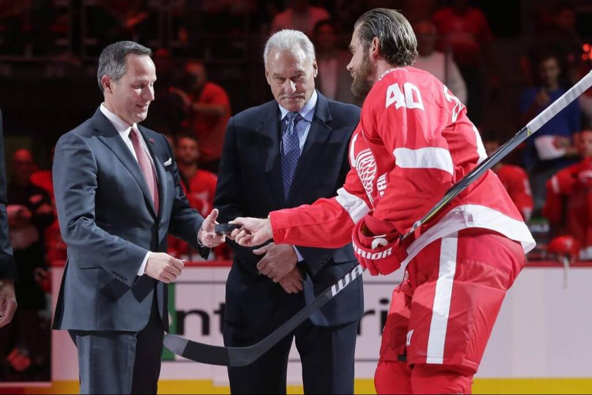 Detroit Red Wings CEO Christopher Ilitch, left, is handed the ceremonial first puck by Henrik Zetterberg (40) as Mickey Redmond, center, looks on.