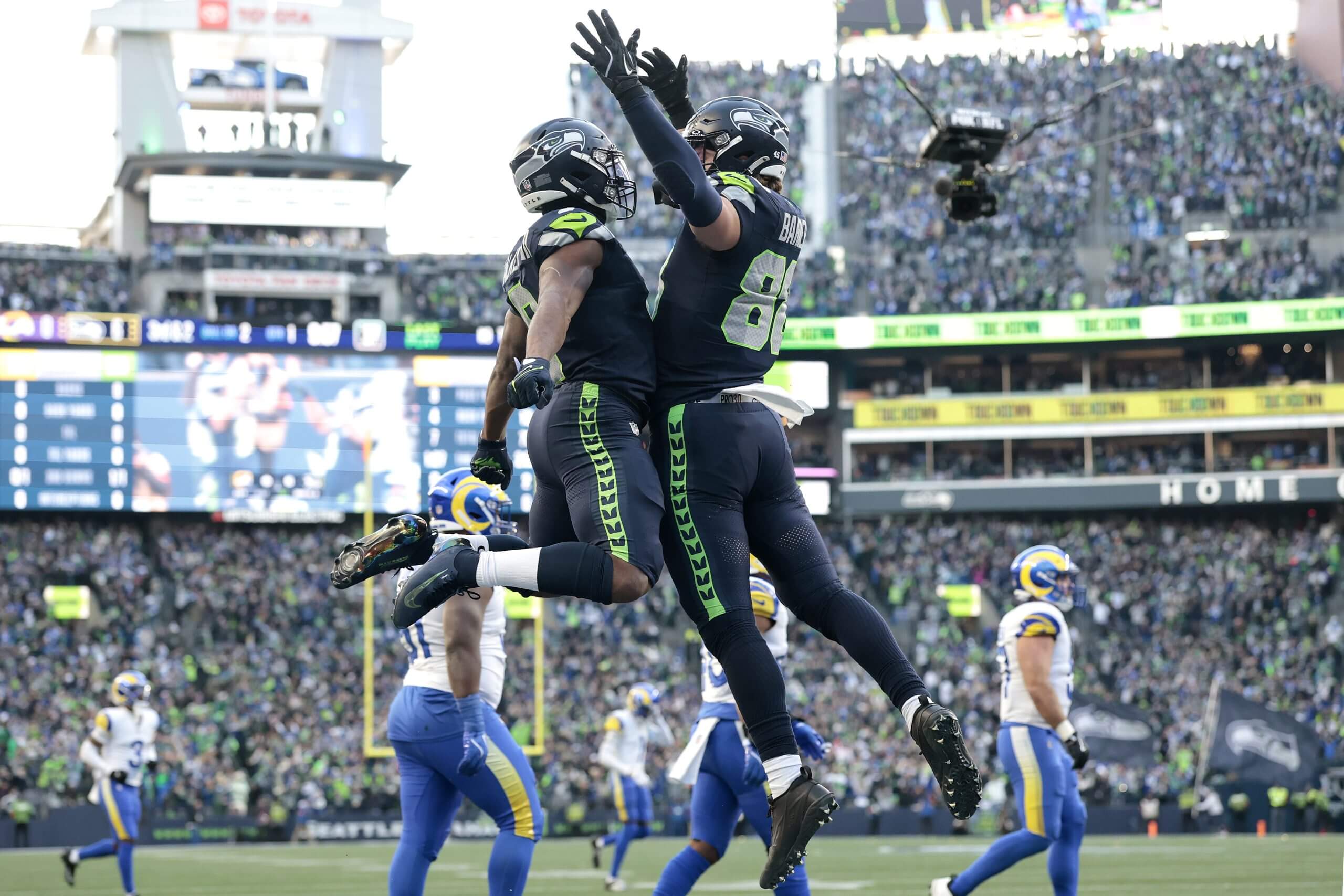 Kenneth Walker and A.J. Barner, wearing all navy uniforms and helmets, leap high and bump chests, with Barner on the right raising his hands over his head, as Rams players walk off the field in the background.